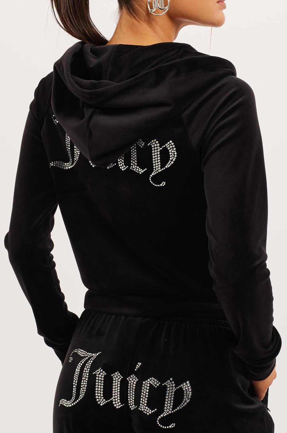 Woman wearing a Black velour Juicy Couture tracksuit with Juicy diamanté branding on the back of the hoodie and the backside of the bottoms